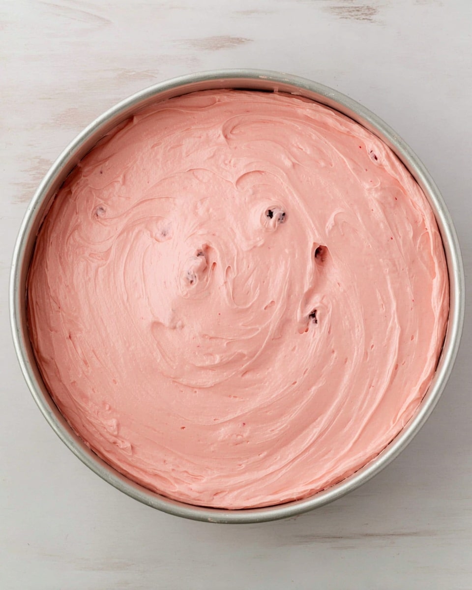 A single round layer of smooth, light pink creamy mixture fills a silver baking pan, with a few small dark spots scattered within the creamy surface. The texture looks soft and whipped, covering the pan evenly and reaching the edges with a few slight swirls on top. The pan rests on a white marbled surface photo taken with an iphone --ar 4:5 --v 7