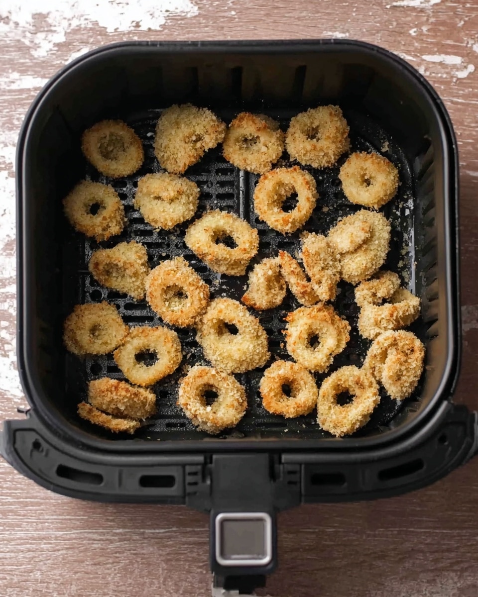 The image shows a black air fryer basket filled with small pieces of food that look like rings and chunks covered in a light golden crumb coating. The coated pieces are spread out evenly inside the basket in about four rows, with some pieces having a round ring shape and others irregular shapes, all with a rough, baked texture. The basket is placed on a wood textured surface, but for your prompt, imagine it on a white marbled texture. photo taken with an iphone --ar 4:5 --v 7