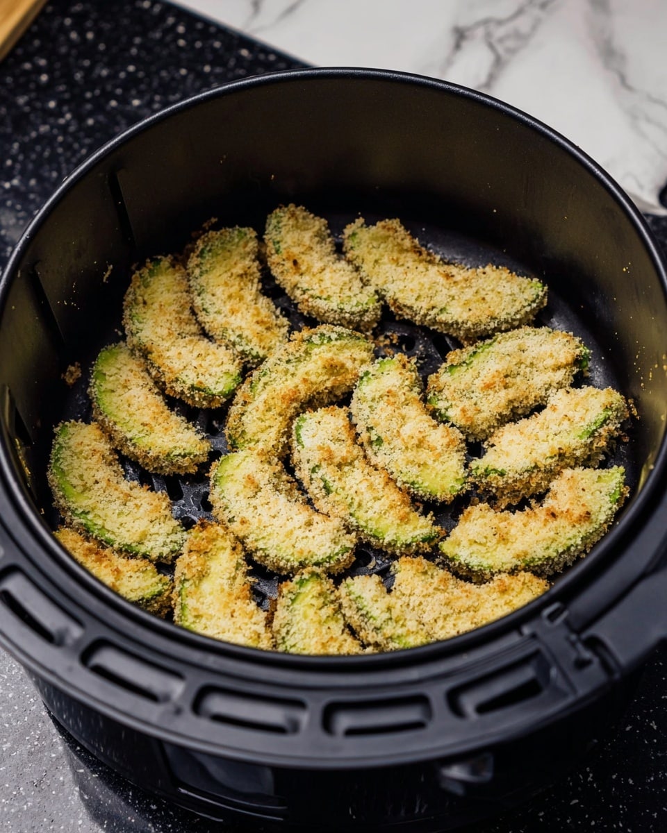 The image shows a black air fryer basket filled with two neat layers of breaded avocado slices. The avocado slices are arranged in a circular pattern with a light golden crumb coating mixed with green avocado visible underneath. The crust texture looks crunchy and evenly spread on each slice. The basket is placed on a black countertop with a white marbled texture in the background. On the side, there is a block of mozzarella cheese partially visible. photo taken with an iphone --ar 4:5 --v 7