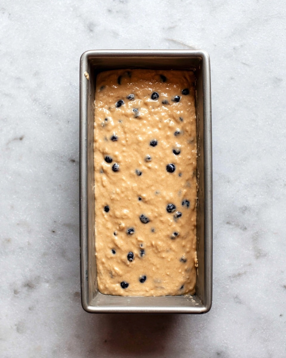 A rectangular metal baking pan filled with a thick, light brown batter that has a slightly lumpy texture, dotted with small dark blue or black round pieces evenly spread throughout. The pan sits on a white marbled surface. Photo taken with an iphone --ar 4:5 --v 7