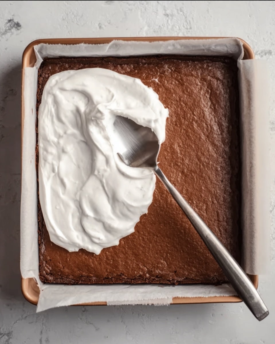 The image shows a square baked chocolate layer with a rich brown color and a slightly cracked surface, placed on a baking tray lined with white parchment paper on a white marbled surface. On the top left portion of the chocolate layer, there is a thick, white creamy layer being spread smoothly with a large metallic spoon, covering about one-third of the top half. The spoon has a shiny reflective surface, angled diagonally with its handle pointing toward the bottom right. The contrast between the matte brown chocolate base and the glossy white cream creates a clear visual divide. Photo taken with an iphone --ar 4:5 --v 7