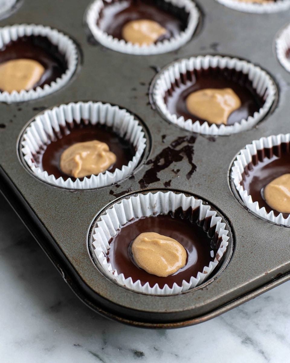 The image shows a close-up of a dark metal muffin tray with six white cupcake liners inside the cups. Each liner has a layer of smooth, dark chocolate coating lining the inside surface of the paper, creating a glossy dark brown base. On top of the chocolate layer, there is a dollop of thick, light brown batter or filling placed in the center of each liner. The tray sits on a white marbled surface, and some spatters of chocolate are visible on the tray edges. photo taken with an iphone --ar 4:5 --v 7