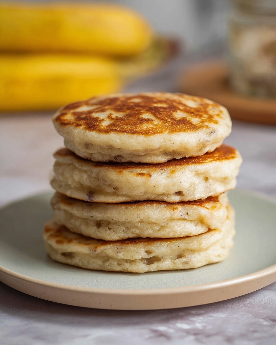 A stack of four thick pancakes sits in the center of a white plate with a soft matte finish. Each pancake is golden brown on top with a slightly uneven, fluffy texture visible on the sides, showing some air bubbles and a light beige color with hints of darker spots. The pancakes are round but not perfectly shaped, giving a homemade look. The background is softly blurred, showing a bunch of yellow bananas and a glass jar on a white marbled surface, giving a cozy kitchen feel. photo taken with an iphone --ar 4:5 --v 7