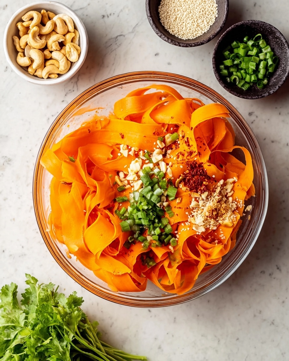 A clear glass bowl filled with thin, wide orange carrot ribbons arranged in loose curls and folds. On top of the carrot ribbons, there are small piles of chopped green onions, minced garlic, white sesame seeds, and a sprinkle of red chili flakes in separate sections. Around the bowl on a white marbled surface, there is a small white bowl with cashew nuts, another small white bowl holding chopped green onions, a small dark bowl containing more white sesame seeds, and a small bunch of fresh green herbs placed near the bottom right. The photo is taken with an iphone --ar 4:5 --v 7