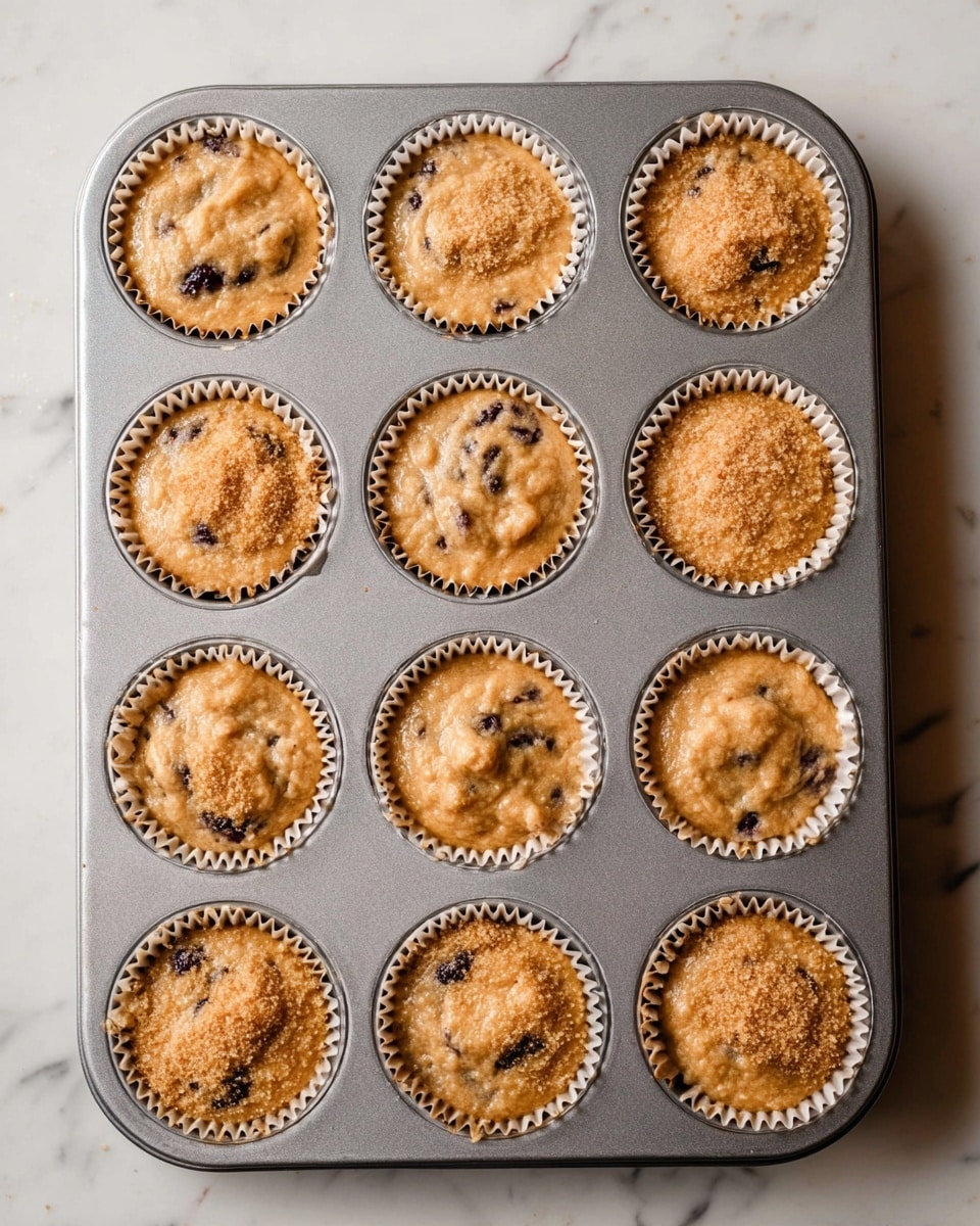A metal muffin tray holds 12 cupcake liners filled with light brown batter that has a smooth, slightly bubbly texture. Darker spots of fruit or chocolate are visible in each cupcake, blending with the batter. The tops have a sprinkle of light brown sugar, creating a grainy layer over the batter. The tray rests on a white marbled surface, and the image is taken from above, showing the full tray and part of the surface. photo taken with an iphone --ar 4:5 --v 7