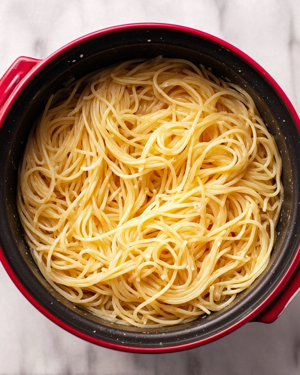 The image shows a close-up of cooked spaghetti noodles inside a black pot with a red outer side. The spaghetti is light yellow, shiny, and slightly tangled, sitting in a thin layer of liquid at the bottom of the pot. The texture of the noodles is smooth and soft, filling the pot evenly without any other ingredients visible. The background is a white marbled texture. photo taken with an iphone --ar 4:5 --v 7