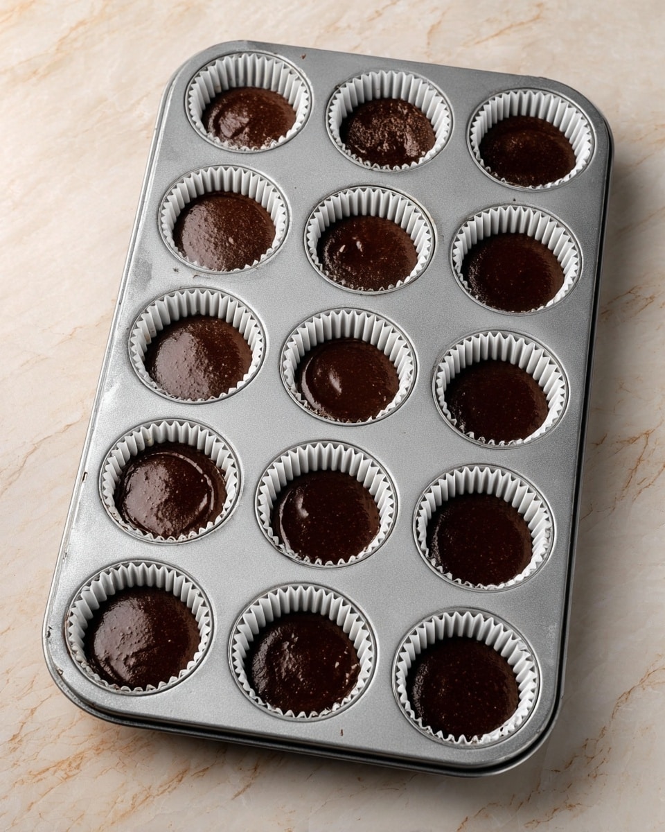 A metal muffin tray with twelve spaces holds chocolate cupcake batter in white paper liners. Each liner is filled about three-quarters full with dark, smooth, and shiny chocolate batter. The tray rests on a white marbled surface, and the batter is evenly spread inside each liner, some showing a slightly glossy wet texture on top. Photo taken with an iphone --ar 4:5 --v 7