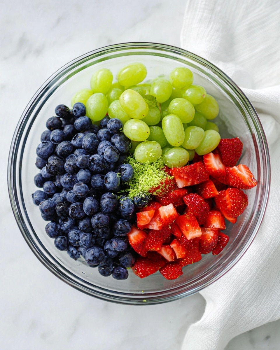 A clear glass bowl on a white marbled surface holds three main sections of fresh fruit: deep blue blueberries clustered tightly on the left, bright green grapes grouped on the top right, and chopped red strawberries filling the bottom right. In the center, there is a small amount of green zest topping the fruits, adding texture and color contrast. A white cloth is placed casually near the bowl. The lighting is bright and natural, highlighting the fresh and juicy look of the fruits. Photo taken with an iphone --ar 4:5 --v 7