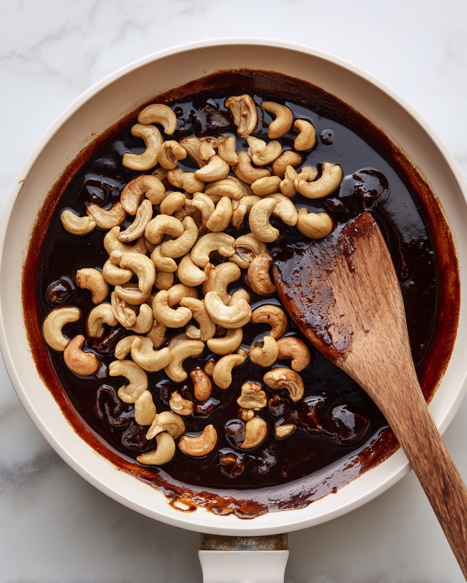 A white pan is filled with small, glossy, dark brown pieces of food mixed with a thick sauce, and topped with a large handful of light beige cashew nuts. A wooden spoon rests inside the pan, partially covered by the cashews and sauce, showing some sauce smeared on its surface. The pan sits on a white marbled surface. Photo taken with an iphone --ar 4:5 --v 7