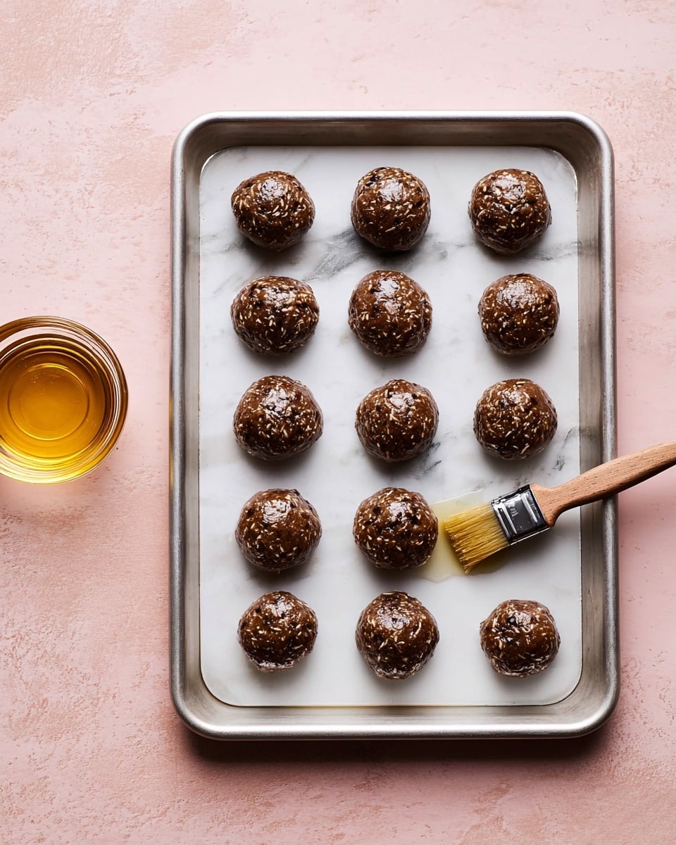 An overhead view of a silver baking tray filled with five rows of five round brown cookie dough balls arranged evenly. Each dough ball looks textured with small bits inside, slightly shiny on top due to a thin coat of glaze being brushed on by a small brush with a light wooden handle. To the left of the tray, there is a small glass container filled with golden liquid, all placed on a white marbled surface with a soft pink surrounding. Photo taken with an iphone --ar 4:5 --v 7