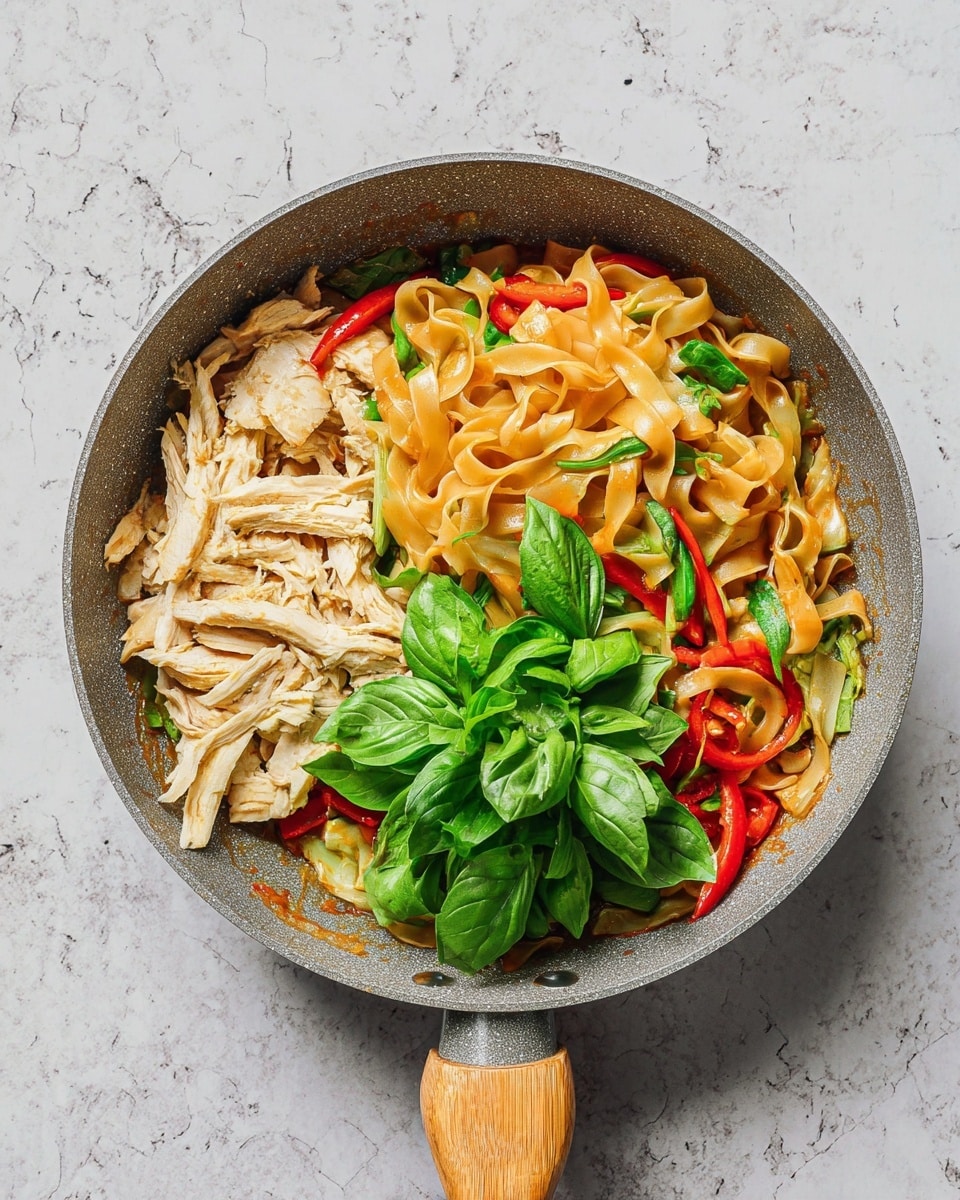 A gray frying pan with a wooden handle holds a colorful dish. At the bottom, wide, flat, light brown noodles mix with green vegetables and red chili slices. On the left side, there are pale strips of cooked chicken layered above the noodles. On the right side, a small pile of fresh green basil leaves rests on top of the noodles. The pan sits on a white marbled surface. photo taken with an iphone --ar 4:5 --v 7