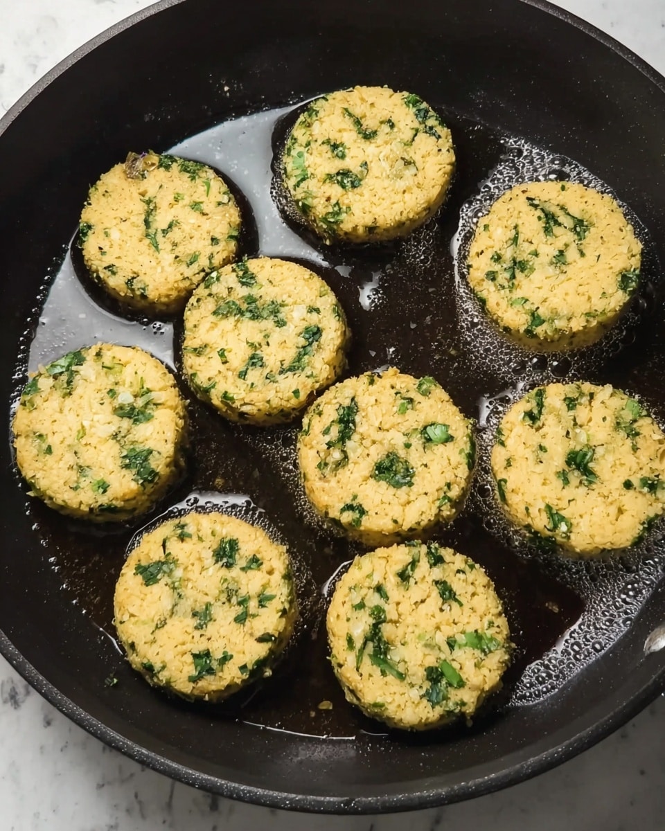 The image shows a black pan with nine round patties cooking in oil. Each patty is pale yellow with small green bits mixed throughout, giving them a textured and speckled look. The patties are closely placed in the pan, their edges slightly browned, and the oil around them reflects light, creating a shiny surface. The background is a white marbled texture. photo taken with an iphone --ar 4:5 --v 7