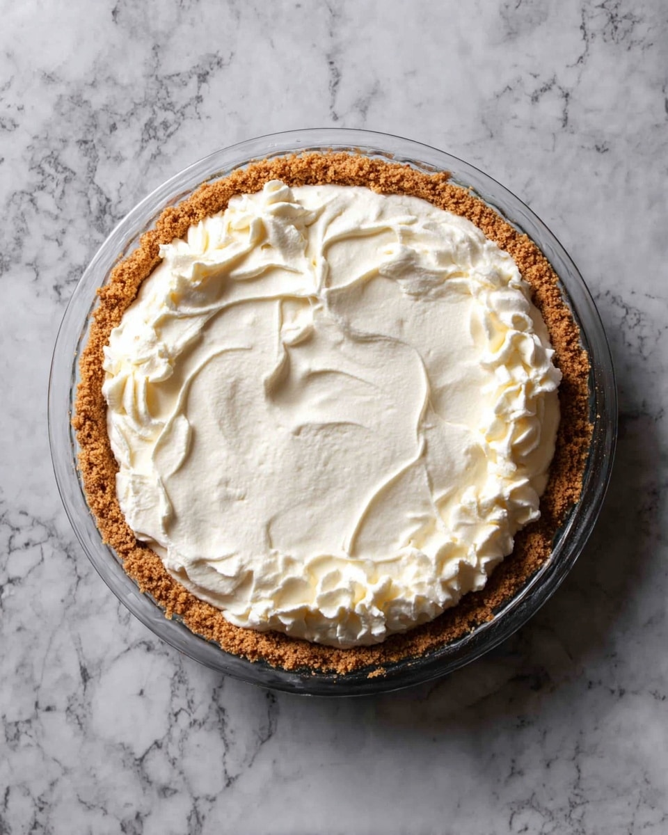 A round pie in a clear glass pie dish with a golden brown crumb crust forming the base and sides. The top layer is thick, white, and creamy with a smooth center and waves of whipped cream swirled around the edge. The pie is placed on a white marbled surface, showing a neat and inviting texture. photo taken with an iphone --ar 4:5 --v 7