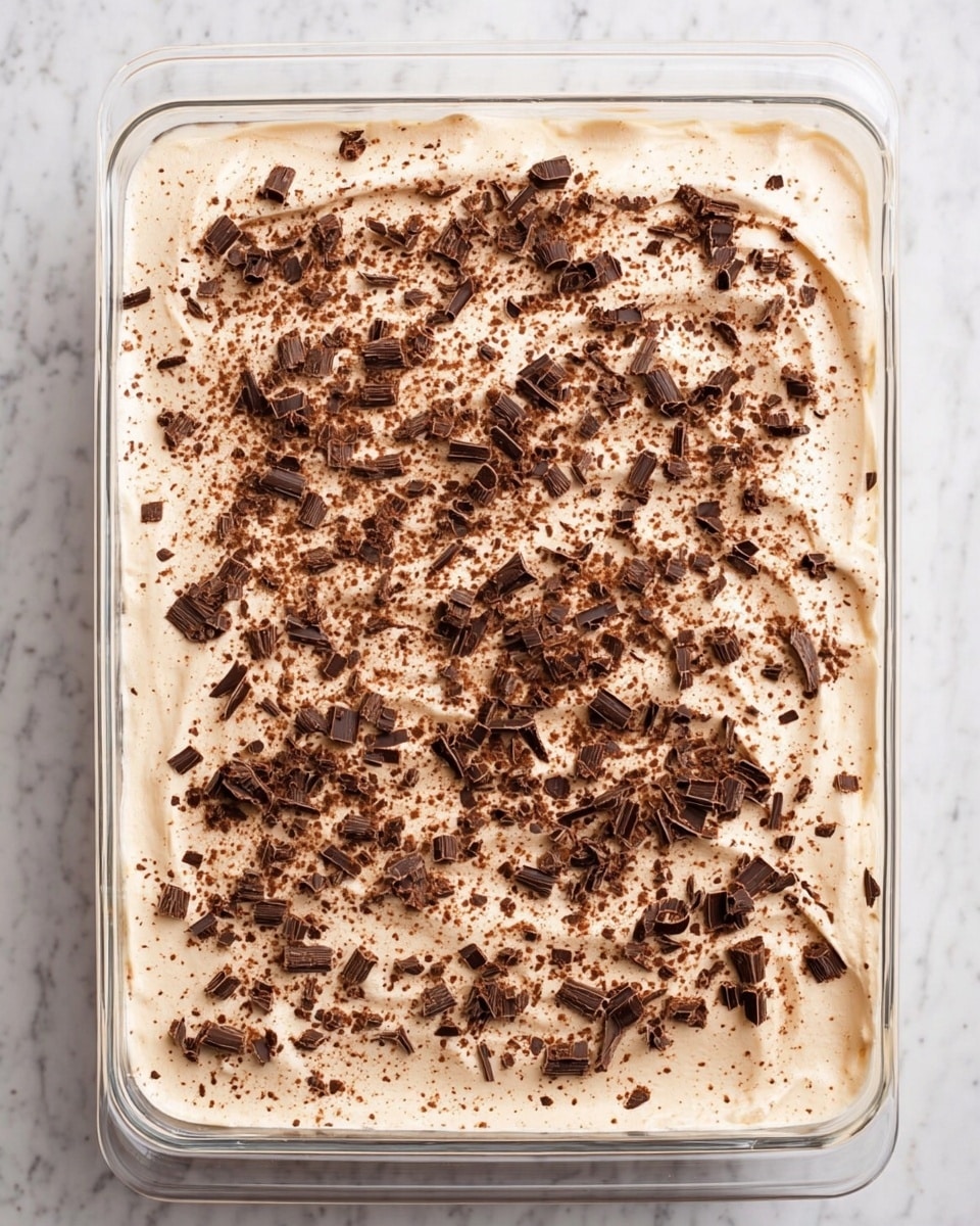 A clear glass rectangular dish holds a layered dessert with a smooth, creamy light beige top layer spread evenly. Dark brown chocolate shavings and small chocolate curls are scattered across the entire surface, adding texture and contrast. The dish rests on a white marbled texture surface, and the image is taken from above, showing the full top layer and the clear sides of the dish. photo taken with an iphone --ar 4:5 --v 7