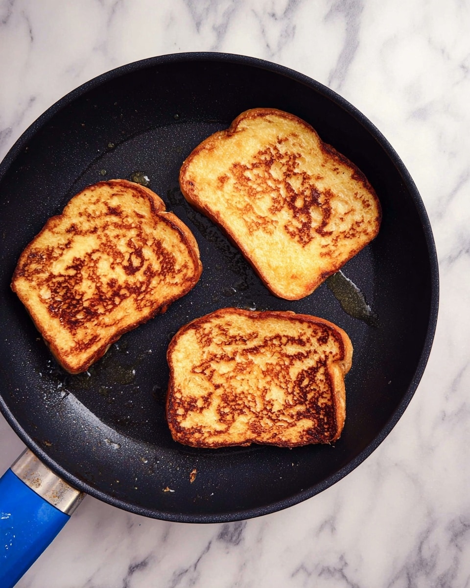 Three slices of golden brown French toast are in a black frying pan with a blue handle. Each toast slice shows a slightly different pattern of browning, with crispy edges and soft, spongy centers. The pan sits on a white marble surface, giving a clean and bright look. Small drops of oil glisten slightly on the pan around the toast. Photo taken with an iphone --ar 4:5 --v 7