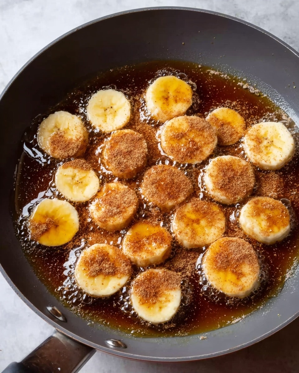 A gray frying pan filled with golden syrup bubbling around several banana slices. The banana slices are spread evenly across the pan with some slices topped with a layer of light brown cinnamon powder. The bananas are a pale yellow color with a soft texture, and the syrup is dark amber, glossy, and thick. The pan rests on a white marbled surface. Photo taken with an iphone --ar 4:5 --v 7