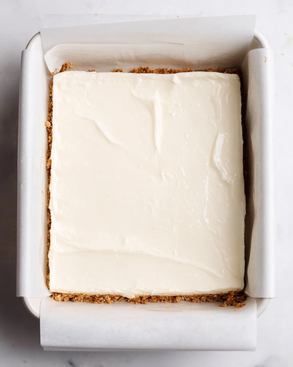 A square white baking dish with two pieces of white parchment paper under the edges holds a dessert with two visible layers; the bottom layer is light brown and crumbly with some crumbs around the edges, and the top layer is smooth, thick, and creamy white, spread evenly across the surface. The dish is placed on a white marbled background. photo taken with an iphone --ar 4:5 --v 7