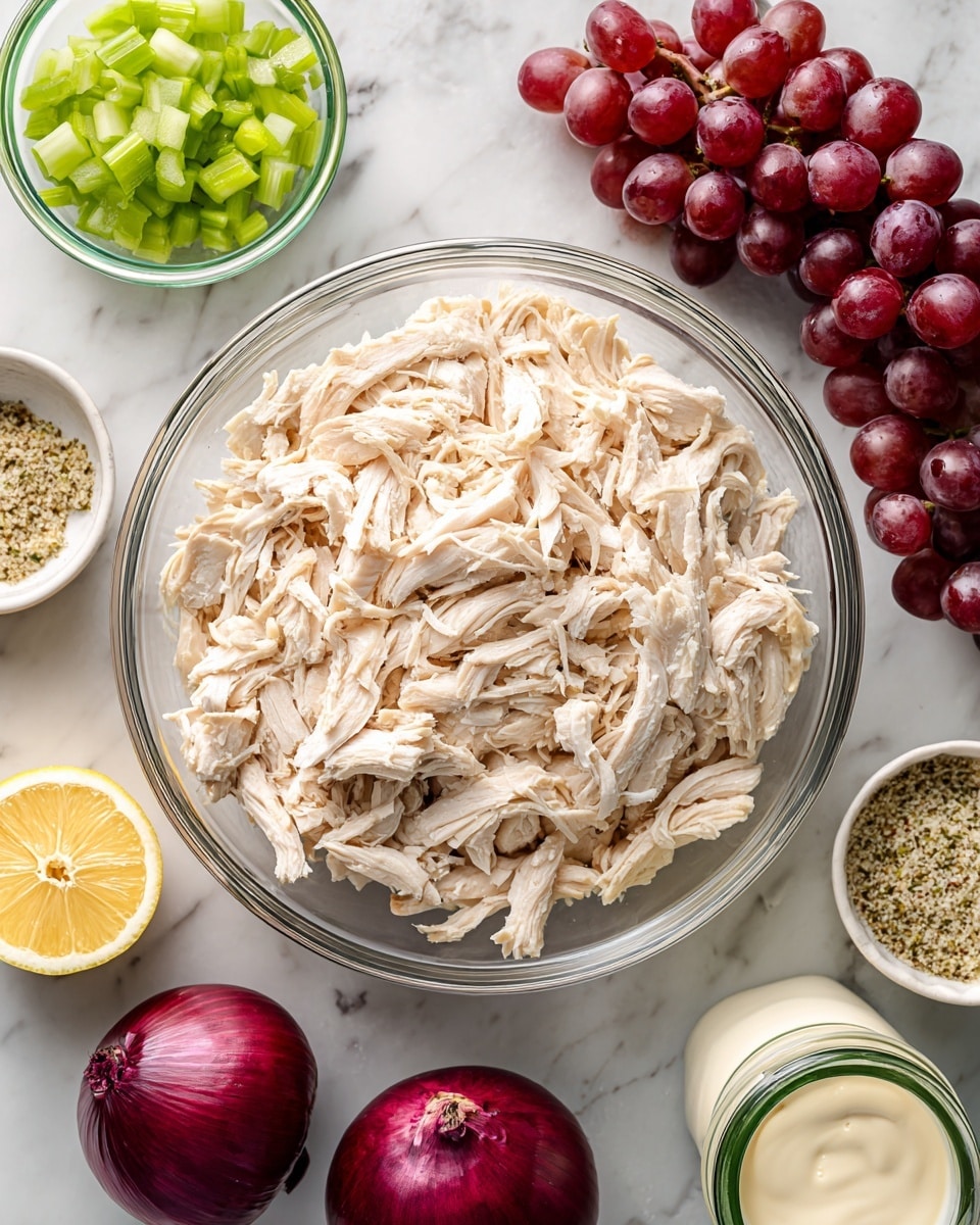 A large clear glass bowl filled with shredded white chicken sits in the middle on a white marbled surface. Above the bowl to the left is a smaller clear glass bowl with small green celery pieces. Next to it on the right is a bunch of red grapes. To the right of the chicken bowl is a small round white bowl holding a mix of everything bagel seasoning. Below the chicken bowl on the left is a lemon, shown in half with bright yellow color and juicy texture. Near the bottom center is a whole red onion with deep purple skin and root end visible. On the surface to the right is a white jar of mayo with a green lid. photo taken with an iphone --ar 4:5 --v 7