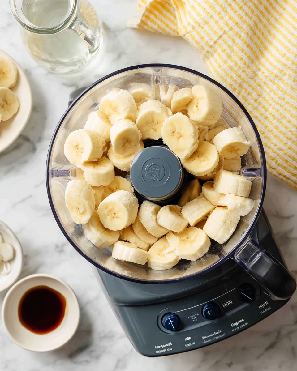 A clear food processor bowl filled with two layers of sliced yellow bananas, each slice thick and smooth with a soft texture. The food processor base is dark grey with blue buttons labeled