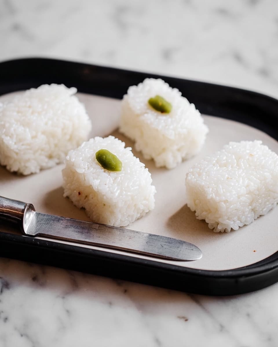 The image shows five pieces of sushi rice shaped in small rectangular blocks, placed on a white plate with a black interior. Each rice block is white with a slightly shiny, sticky texture. One piece has a small green topping in the center. The plate is on a white marbled surface, and a silver knife is partially visible next to the plate. The photo taken with an iphone --ar 4:5 --v 7