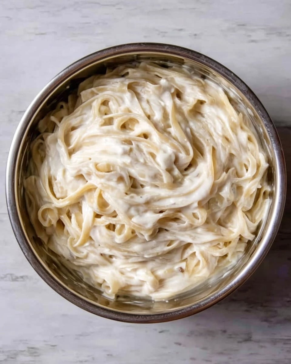 A round metal bowl filled with creamy white pasta ribbons, layered in a slightly messy swirl, showing a smooth, thick sauce coating all the noodles evenly. The sauce has a shiny, silky texture with small bits giving it a rich look. The bowl sits on a white marbled surface, highlighting the pasta's pale color and creamy texture. photo taken with an iphone --ar 4:5 --v 7