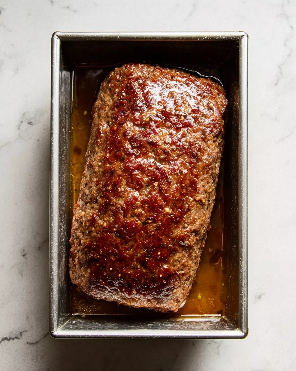 A single large meatloaf with a browned and slightly shiny surface sits inside a rectangular metal baking pan filled with some juices. The texture of the meatloaf is coarse with small bits visible, and the edges reflect light softly. The pan is placed on a white marbled surface, visible around the pan. There are no other objects in the image. photo taken with an iphone --ar 4:5 --v 7