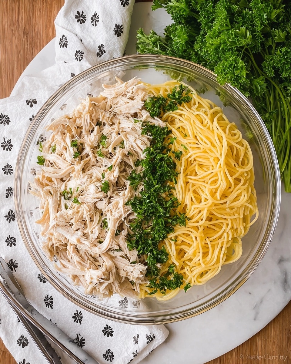 A clear glass bowl sits on a white marbled surface, filled with two main layers side by side: on the left is a pile of shredded light beige cooked chicken, and on the right is a mound of cooked yellow spaghetti noodles. Bright green chopped parsley is sprinkled in a line down the middle, separating the chicken and pasta, with some parsley scattered on top. Near the bowl, there is a bunch of fresh parsley and a white cloth with small black floral patterns resting on the white marbled texture. A pair of metal tongs lies next to the bowl. Photo taken with an iphone --ar 4:5 --v 7