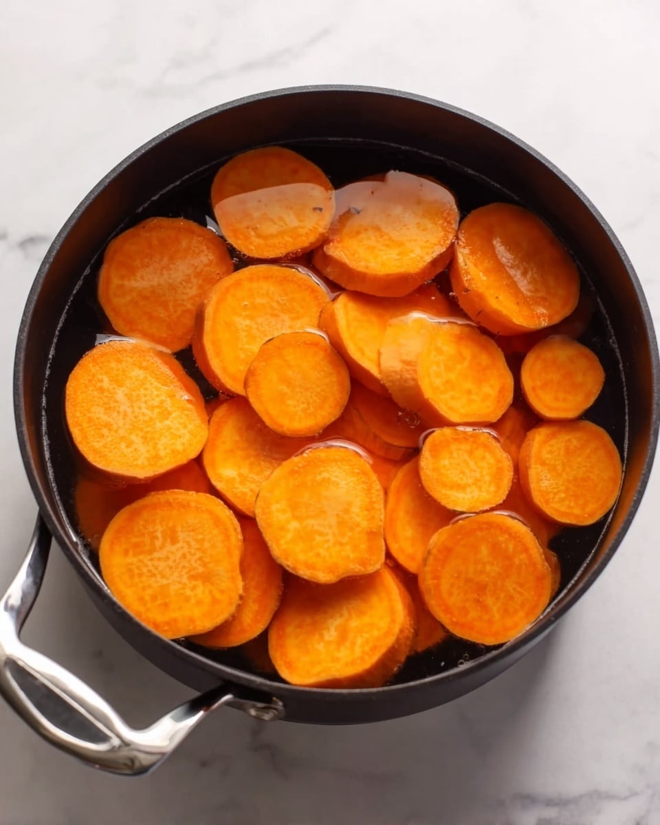 A black cooking pot filled with thick slices of bright orange sweet potatoes, partially submerged in clear water. The sweet potato slices vary slightly in size and have a smooth, soft texture. The pot has a shiny silver handle and is placed on a white marbled surface. The water reflects some light, adding a slight shine to the sweet potato pieces. photo taken with an iphone --ar 4:5 --v 7