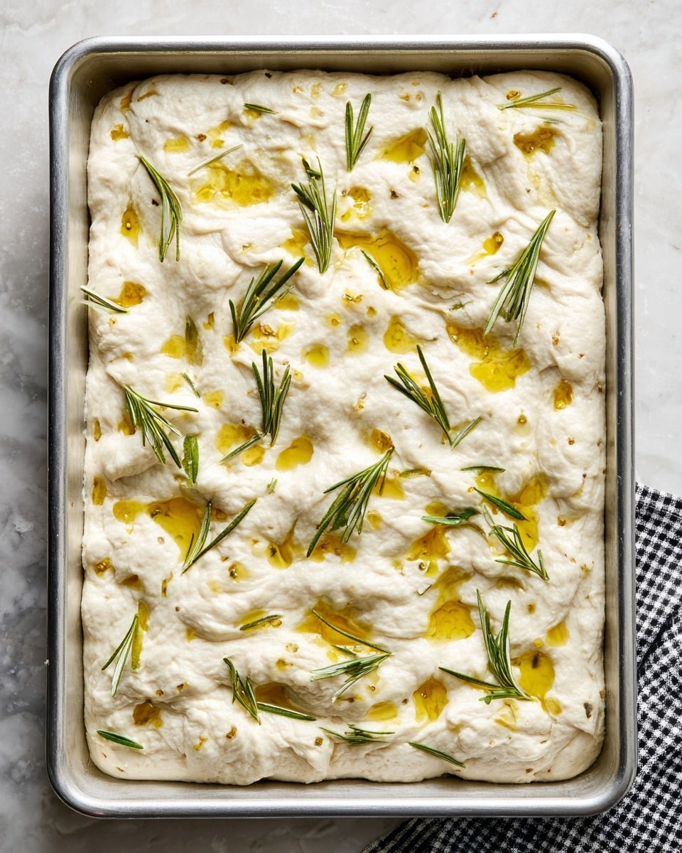 A rectangular silver baking tray filled with thick, fluffy dough that is puffy and bumpy on the top layer. The dough is white with a soft, airy texture and is dotted with small pools of golden olive oil that shine on the surface. Bright green rosemary leaves are scattered evenly across the top, adding a fresh and natural touch. The tray is placed on a white marbled surface with a black and white checkered cloth peeking from the bottom left corner. Photo taken with an iphone --ar 4:5 --v 7