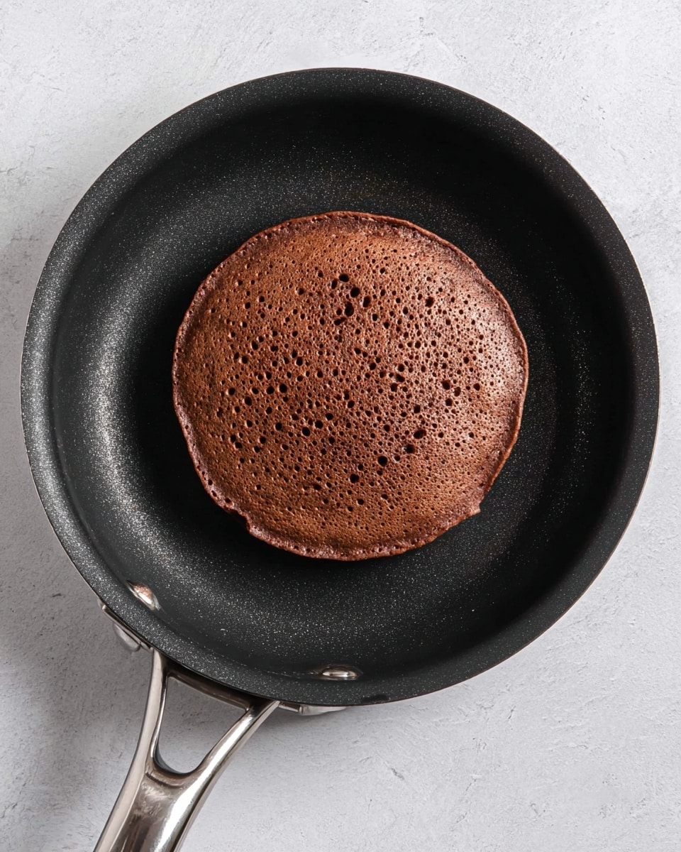 A single round chocolate pancake with a slightly textured and bubbled surface sits in the center of a black non-stick frying pan, which has a silver handle extending out to the left. The pancake is a rich dark brown color with small holes and a slightly uneven edge. The black pan contrasts with the pancake and rests on a white marbled surface. photo taken with an iphone --ar 4:5 --v 7