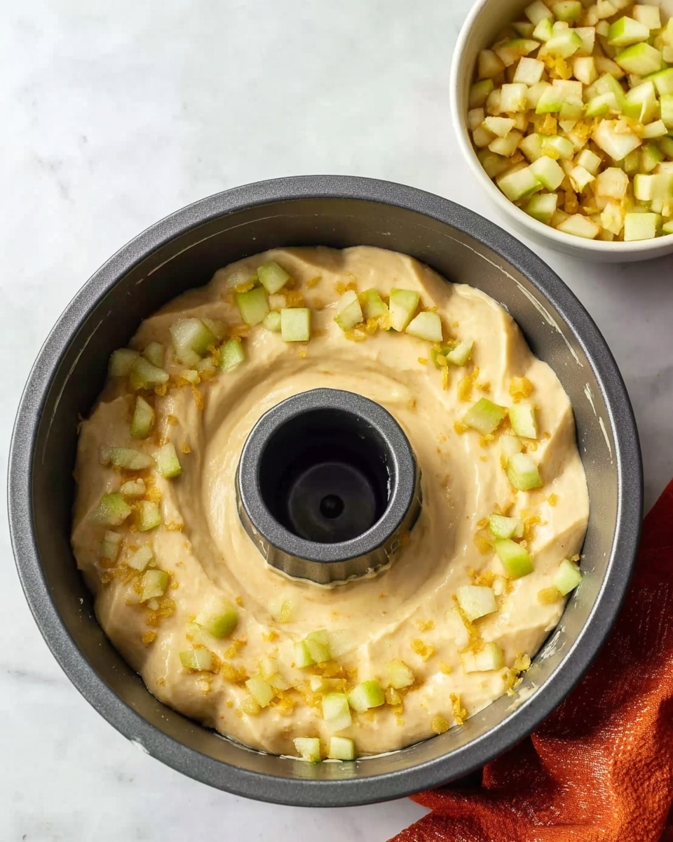 A dark gray bundt pan filled with a thick, pale yellow batter spread evenly along the bottom, with a hollow center visible. On top of the batter, small, light green and yellow diced fruit pieces are scattered evenly, adding texture and color contrast. Nearby, a white bowl filled with the same diced fruit sits on a white marbled surface, with an orange cloth partially visible in the lower right corner. The lighting is soft and natural, showing slight shine on the glossy batter surface. photo taken with an iphone --ar 4:5 --v 7