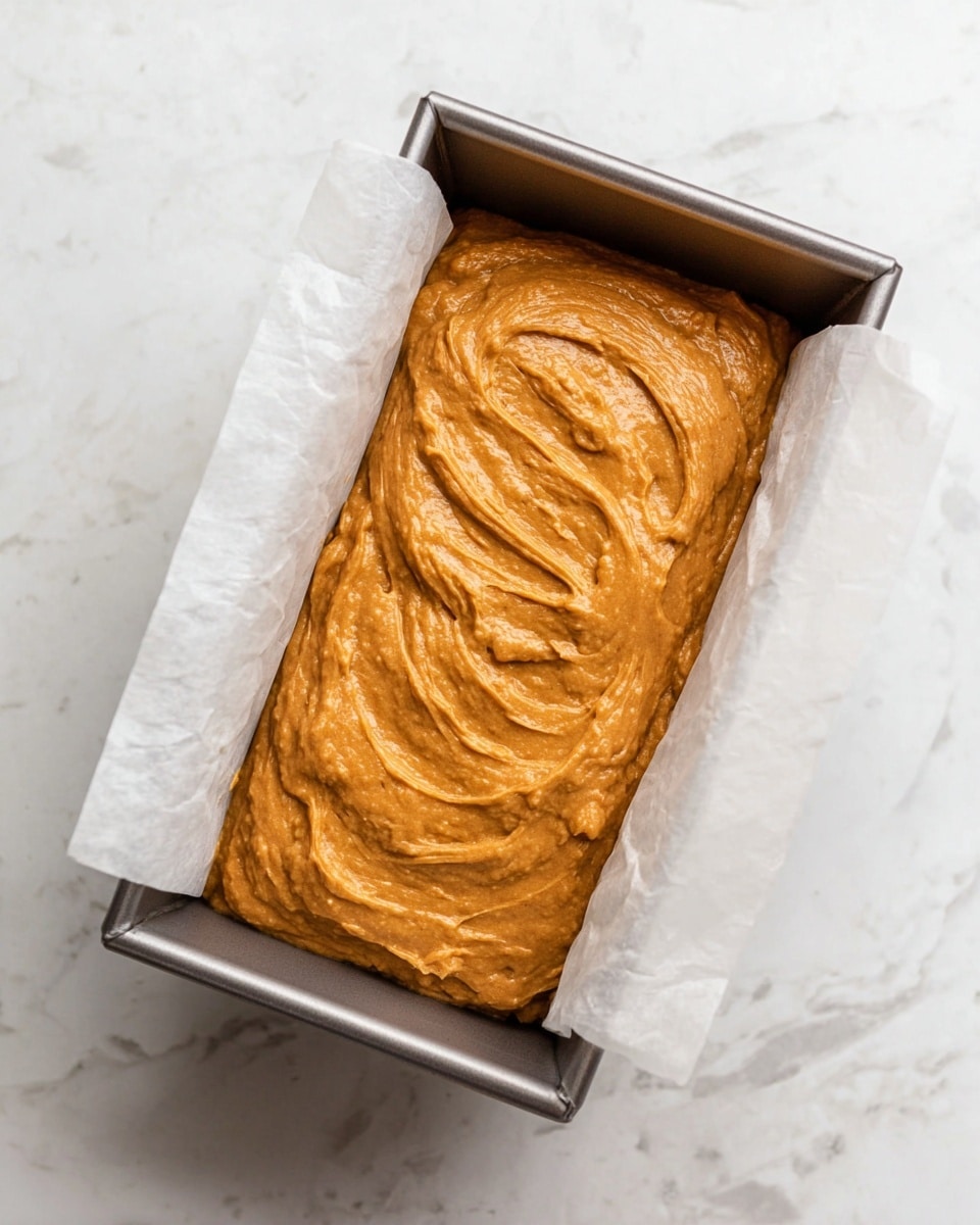 A metal bread pan lined with white parchment paper holds two layers of orange-brown batter that look thick and smooth, with swirled textures on the top layer. The pan is placed on a white marbled surface that adds a clean and bright background to the scene. The batter fills most of the pan, showing a soft and creamy consistency ready for baking. Photo taken with an iphone --ar 4:5 --v 7