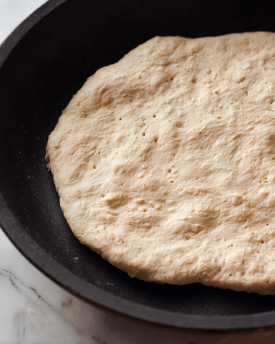 A close-up view of a single flat, round piece of light beige dough with a slightly uneven and rustic texture, resting in a dark pan. The dough has small air bubbles and a few darker specks scattered across its surface, with edges that are slightly thicker and irregular in shape. The pan contrasts with the pale dough, highlighting its soft and smooth appearance. The background shows a glimpse of a white marbled surface. photo taken with an iphone --ar 4:5 --v 7