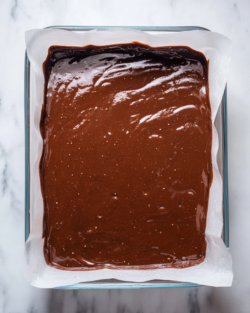 A clear rectangular glass dish lined with white parchment paper holds a single thick layer of smooth, glossy dark brown chocolate batter with small light spots and bubbles visible on the surface. The batter is evenly spread to fill the dish, showing some slight swirls and ridges. The dish is set on a white marbled surface. photo taken with an iphone --ar 4:5 --v 7