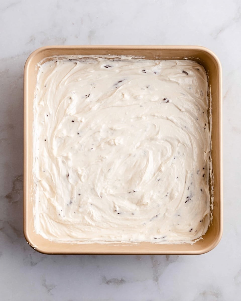 The image shows a square beige baking dish filled with a single thick layer of creamy white mixture that has small dark specks scattered throughout. The mixture is spread smoothly, with some gentle swirls and uneven edges where it meets the sides of the dish. The dish sits on a white marbled surface. photo taken with an iphone --ar 4:5 --v 7