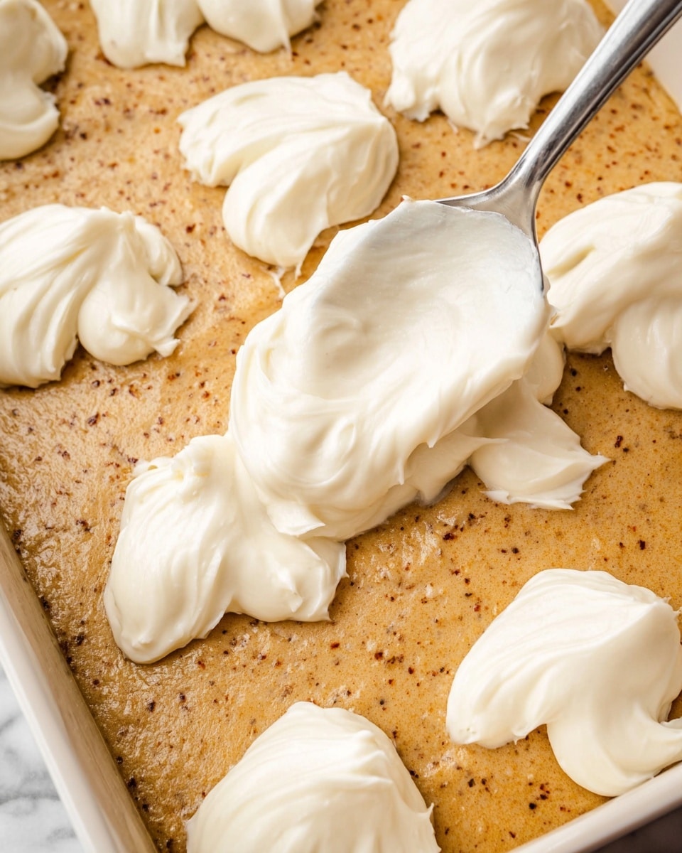 A close-up image shows a white spoon spreading dollops of smooth, creamy white frosting over a thick, light brown batter with specks of darker brown throughout, all inside a white baking pan; the batter looks soft and dense, while the frosting forms rounded, fluffy mounds scattered evenly on top; the scene is set on a white marbled surface. photo taken with an iphone --ar 4:5 --v 7