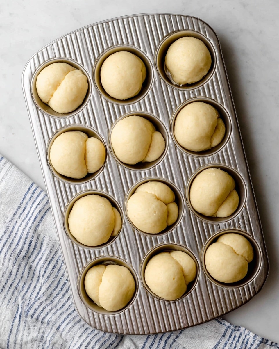 A metal muffin tray filled with 12 round dough pieces, each formed by three small dough balls pressed closely together, creating a smooth, pale yellow surface with a slightly shiny texture, set on a white marbled surface with part of a striped kitchen cloth visible in the lower left corner, the tray's metallic ridges reflect light softly across its surface photo taken with an iphone --ar 4:5 --v 7