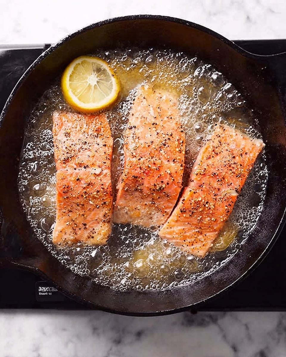 Three pieces of salmon fillet are frying in a black cast iron pan filled with bubbling oil. The salmon has a pinkish-orange color with visible black pepper sprinkled on top. Two lemon wedges, one on the left side and one tucked between two salmon pieces at the top, add a touch of bright yellow. The pan sits on a black stovetop, and the background shows a white marbled surface. The scene looks hot and fresh, capturing the cooking process. photo taken with an iphone --ar 4:5 --v 7