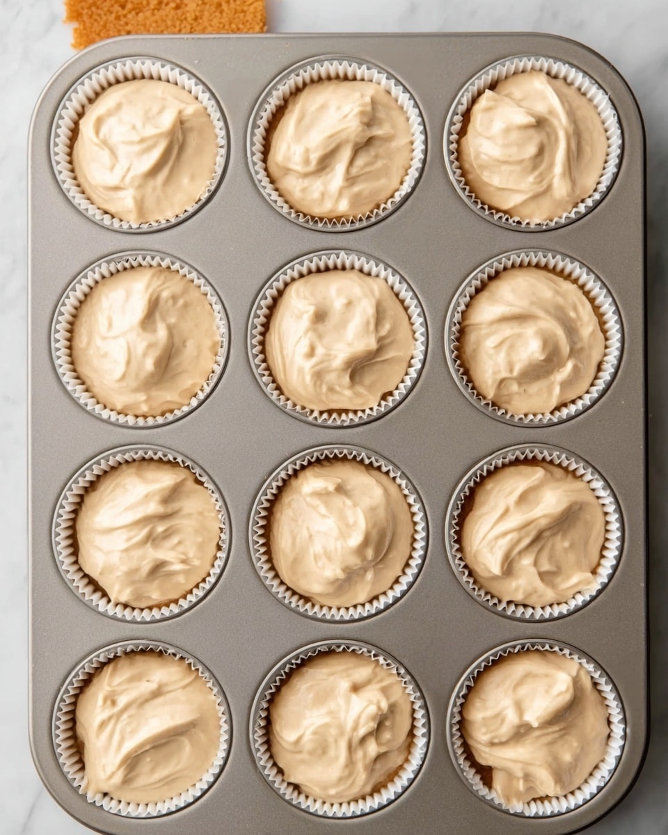 A metal muffin tray with twelve white paper liners filled with light brown batter, each liner neatly holding the same creamy, smooth mixture. The batter in each liner is thick with soft swirls and small air bubbles, slightly domed but not overflowing. The tray rests on a white marbled surface, and one liner on the top left is partially covered by something orange. photo taken with an iphone --ar 4:5 --v 7
