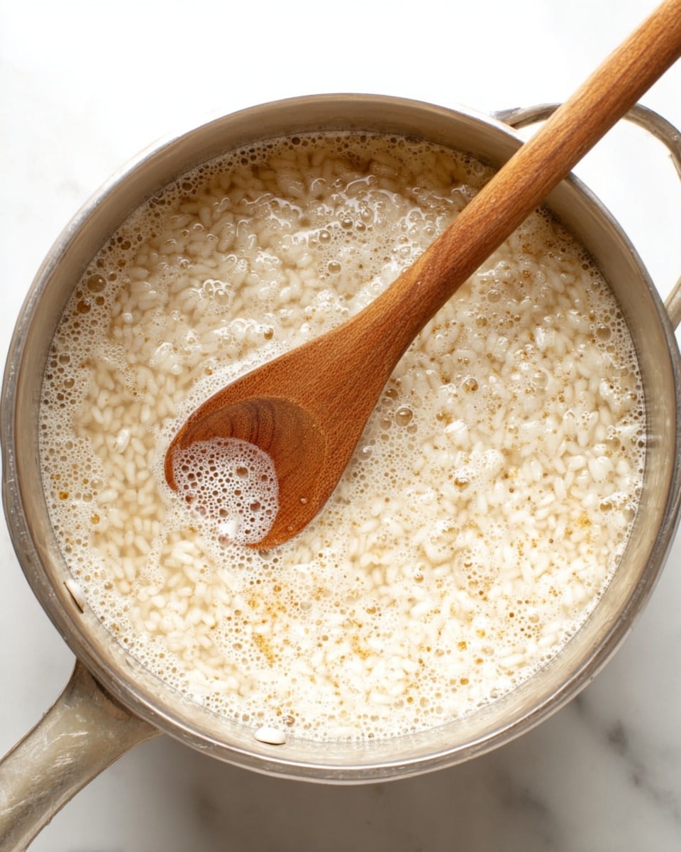 A top view of a silver pot filled with creamy, light beige risotto rice that has small bubbles forming on the surface, showing it is cooking. A wooden spoon with a smooth texture is partially submerged, stirring the mixture. The background is a white marbled texture. Photo taken with an iphone --ar 4:5 --v 7
