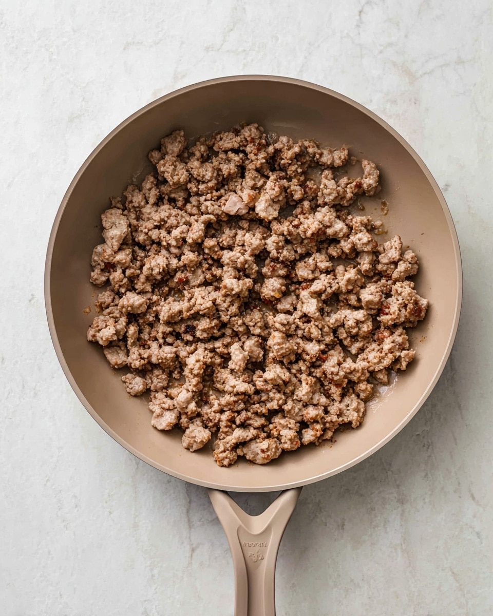 A beige frying pan with cooked ground meat inside fills the image, positioned on a white marbled surface. The ground meat is broken into small to medium chunks, showing a cooked texture with a mix of light and darker brown colors. The pan handle points downward and has a smooth, matte finish. There are no other items or layers visible in the image. photo taken with an iphone --ar 4:5 --v 7