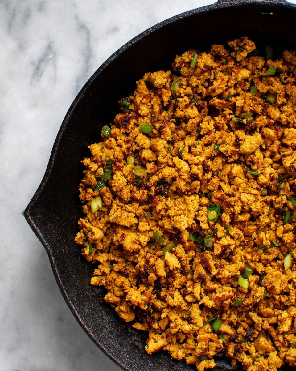 The image shows a black cast iron pan filled with cooked, crumbled textured soya in various shades of golden brown, mixed with small pieces of green herbs and onions. The textured soya has a slightly crispy look, with some darker brown toasted bits and vibrant green flecks scattered evenly throughout. The pan sits on a surface with a white marbled texture, and the focus is on the rich, warm colors and rough texture of the cooked soya inside the pan. photo taken with an iphone --ar 4:5 --v 7