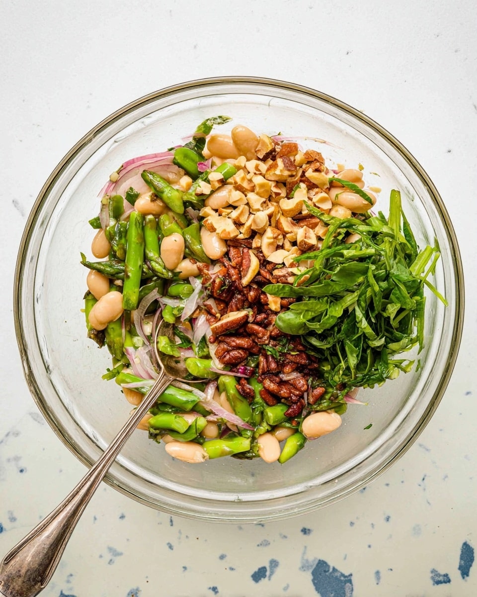 A clear glass bowl contains a colorful salad with multiple layers: at the bottom are light beige beans mixed with bright green asparagus pieces, thin pale pink onion rings, and small chopped dark green herbs. On top of these is a layer of chopped brown nuts scattered around, and a small pile of fresh dark green leafy herbs on the right side. A vintage silver spoon rests inside the bowl on the left. The bowl sits on a white marbled surface with small blue specks. photo taken with an iphone --ar 4:5 --v 7