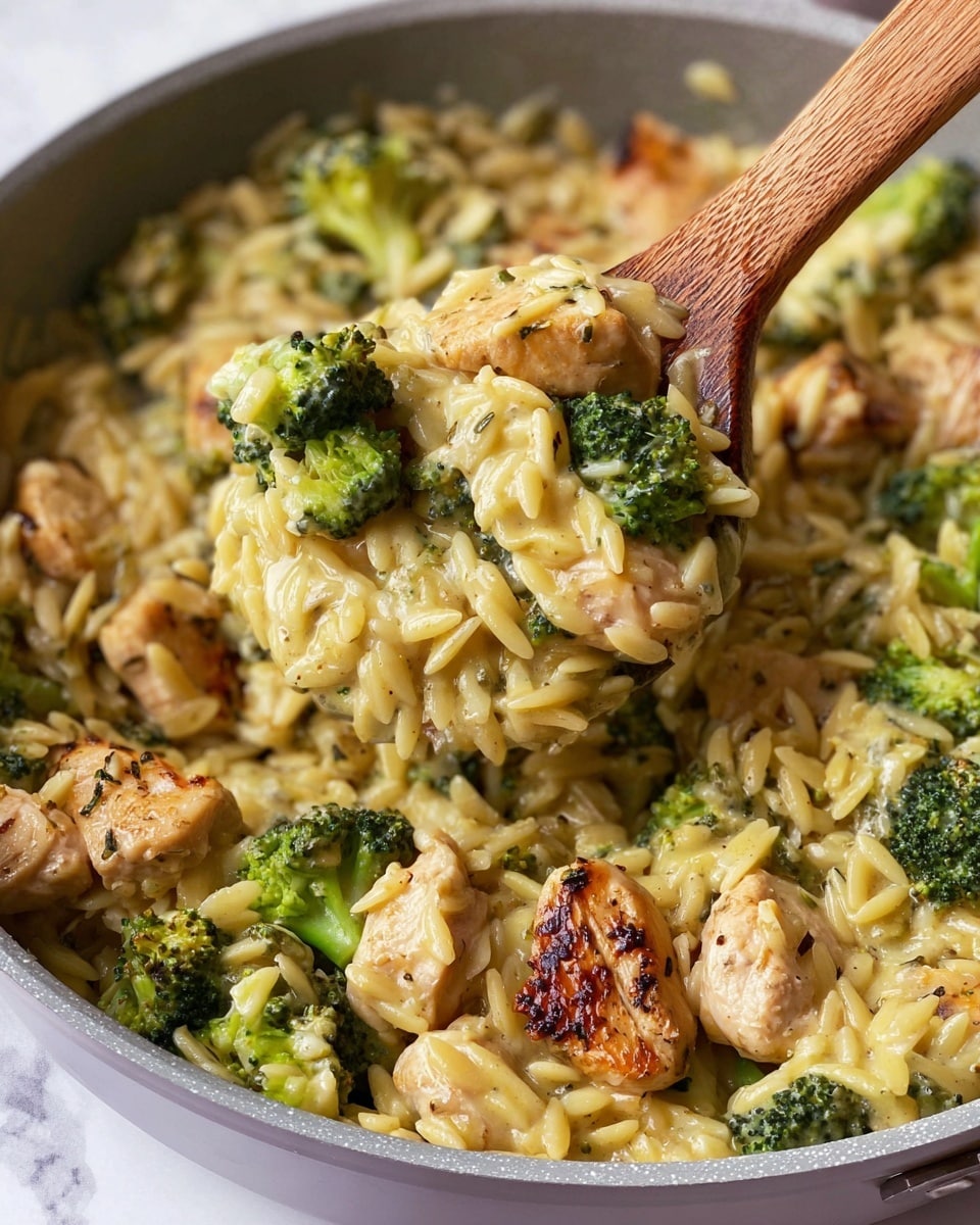 A close-up view of a creamy orzo dish in a light gray pan, showing pieces of cooked chicken with light brown grill marks mixed with bright green broccoli florets and shiny, tender orzo pasta in a thick light yellow sauce. In the foreground, a wooden spoon lifts a generous scoop of the dish, highlighting the mix of soft chicken chunks, green broccoli, and firm orzo. The background is a white marbled texture. Photo taken with an iphone --ar 4:5 --v 7