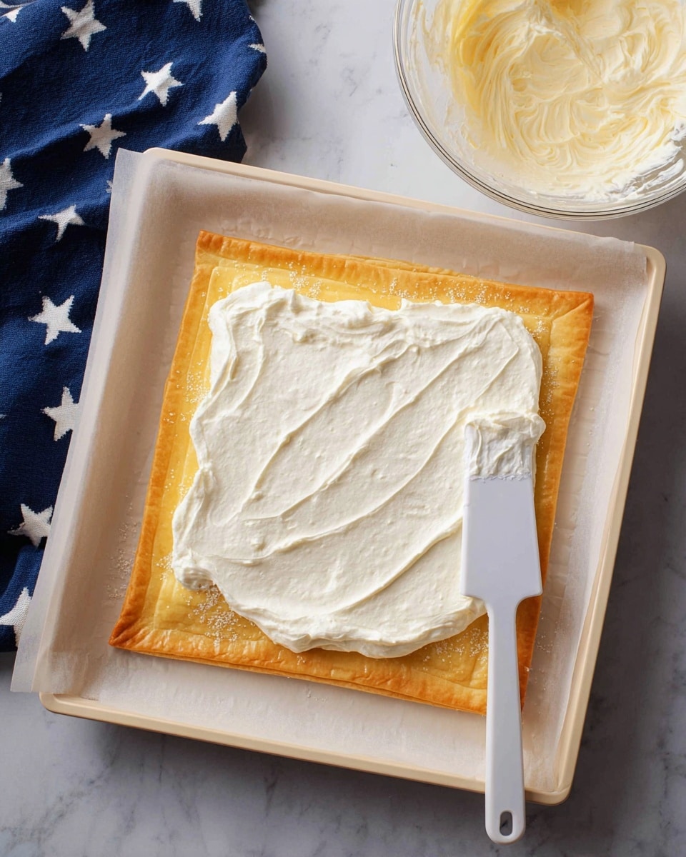 A square, golden-brown puff pastry sheet lies flat on white parchment paper inside a white baking tray. The pastry has a slightly raised, flaky edge and is spread with a thick, smooth layer of white creamy mixture that covers most of its surface. A white spatula rests on the right side, partially coated with the creamy layer. In the upper right corner, a clear bowl filled with more white creamy mixture is visible. The background is a white marbled surface with a navy blue cloth with white stars peeking in at the bottom left corner. photo taken with an iphone --ar 4:5 --v 7