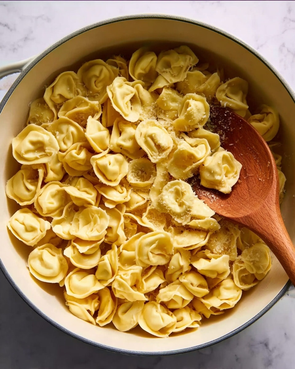 A white round pot filled with many tortellini pasta pieces, light yellow in color, with soft, folded shapes. A wooden spoon with a smooth texture rests inside the pot, partially mixing the pasta. The pot is placed on a white marbled surface that adds a clean, simple background to the image. photo taken with an iphone --ar 4:5 --v 7