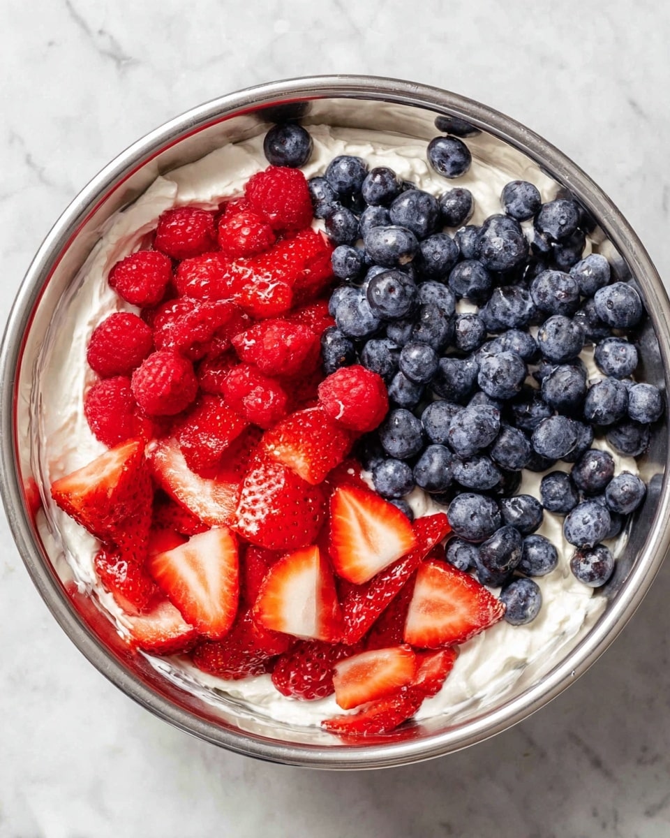 A metal bowl with three parts of fruit on top of a layer of white creamy texture: the top right section has bright red raspberries, the bottom right section shows sliced strawberries with red and white inside, and the left section is filled with whole blue blueberries. The creamy white layer is spread unevenly around the bowl edges, sitting on a white marbled surface. photo taken with an iphone --ar 4:5 --v 7