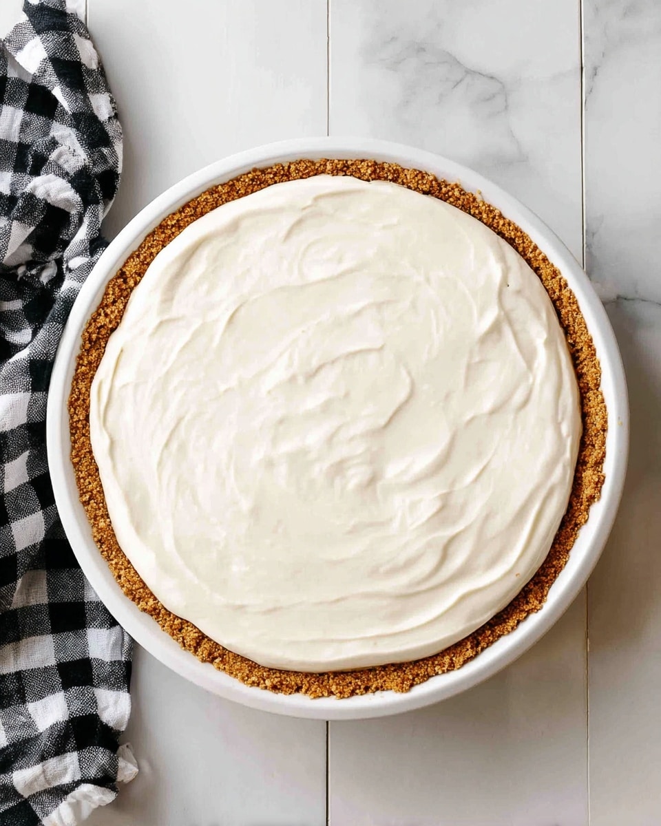 A round pie with two layers is shown in a white pie dish placed on a white marbled texture. The bottom layer is a golden-brown crumbly crust that forms a thick edge around the pie. The top layer is a smooth and creamy white filling spread evenly, with slight swirls visible on the surface. A black and white checkered cloth is partially visible in the bottom left corner. photo taken with an iphone --ar 4:5 --v 7