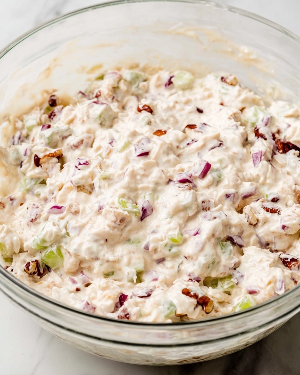 The image shows a close-up of a clear glass bowl filled with a creamy, chunky salad mixture. The creamy layer is off-white with small pieces of red onion, light green celery, and bits of reddish-brown nuts or dried fruit mixed evenly throughout. The texture looks soft and smooth with some more textured, small chunks visible under the creamy coating. The bowl sits on a white marbled surface, and the mixture fills most of the bowl's space. Photo taken with an iphone --ar 4:5 --v 7