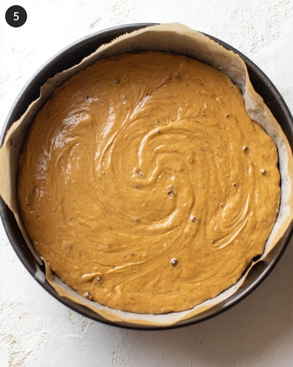 A round baking pan lined with parchment paper holds a single thick layer of smooth, light brown batter with a slightly shiny surface. The batter fills the pan evenly, with some gentle swirls and small bubbles visible on top. The background is a white marbled texture. photo taken with an iphone --ar 4:5 --v 7